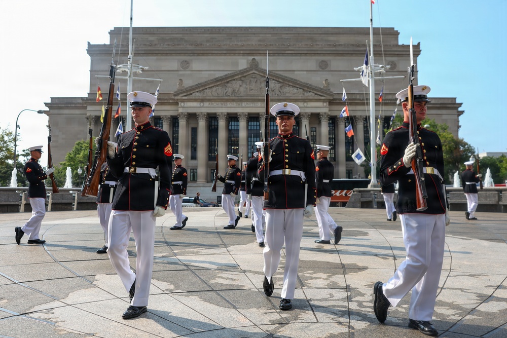 A U.S. Marine Corps ceremonial drill unit is performing a precision marching drill in front of the National Archives Building in Washington, D.C. The Marines are dressed in formal dress blues with white trousers, white gloves, and white covers, carrying rifles. Multiple rows of Marines march in formation across a large plaza area.