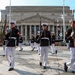 A U.S. Marine Corps ceremonial drill unit is performing a precision marching drill in front of the National Archives Building in Washington, D.C. The Marines are dressed in formal dress blues with white trousers, white gloves, and white covers, carrying rifles. Multiple rows of Marines march in formation across a large plaza area.