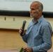 An older man with gray hair and a white beard is speaking into a handheld microphone at what appears to be a public meeting or presentation. He is wearing a blue plaid button-up shirt and holding a small green book or notepad in his other hand. A projection screen with partially visible text is visible in the background.