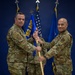 Two military personnel in OCP (Operational Camouflage Pattern) uniforms are participating in a formal guidon-passing ceremony, likely a change of command. They are jointly holding a blue unit guidon bearing the designation '152 ADS' with yellow lettering. A third service member stands in the background holding additional flags.