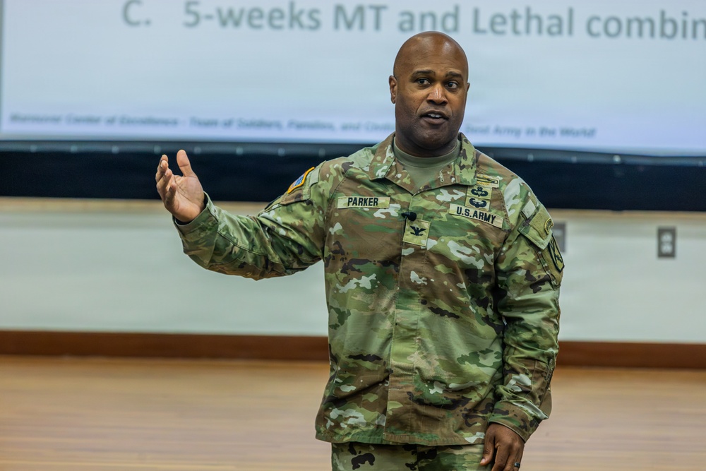 A U.S. Army Colonel named Parker is delivering a presentation or briefing in what appears to be a military training or conference room. He is wearing the Army Combat Uniform (ACU) in OCP (Operational Camouflage Pattern) and gesturing with his right hand while speaking. A projected slide behind him references '5-weeks MT and Lethal combi...' suggesting a training or readiness briefing.