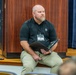 A heavyset, bald man with a beard is seated on the edge of a stage or platform in what appears to be an auditorium or meeting hall. He is wearing a black polo shirt with a logo, khaki pants, and a name badge lanyard, and is holding an open binder or folder. He appears to be attentively listening or observing something off-camera.