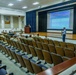 A military briefing or presentation is taking place in an auditorium-style lecture hall. Most attendees are wearing military camouflage uniforms and are seated in rows facing a projection screen displaying a slide titled 'Maneuver Branches Distribution Plan and SMOR Load Projection ASI-U5'. A civilian presenter stands to the right of the screen while another individual is at a podium on the stage.