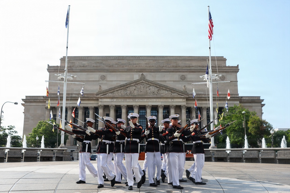 A U.S. Marine Corps Silent Drill Platoon performs a precision rifle drill in front of the National Archives Building in Washington, D.C. The Marines are dressed in their distinctive dress blues with white trousers and white covers, executing synchronized rifle movements. Multiple international and U.S. flags are visible on flagpoles surrounding the neoclassical building.
