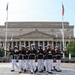 A U.S. Marine Corps Silent Drill Platoon performs a precision rifle drill in front of the National Archives Building in Washington, D.C. The Marines are dressed in their distinctive dress blues with white trousers and white covers, executing synchronized rifle movements. Multiple international and U.S. flags are visible on flagpoles surrounding the neoclassical building.