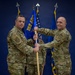 Two military personnel in OCP (Operational Camouflage Pattern) uniforms are conducting a guidon transfer ceremony, likely a change of command or activation ceremony for the 152 AOG (Air Operations Group). Both individuals are gripping a blue guidon staff bearing the unit designation '152 AOG' in gold lettering. A third service member stands in the background holding additional flags.