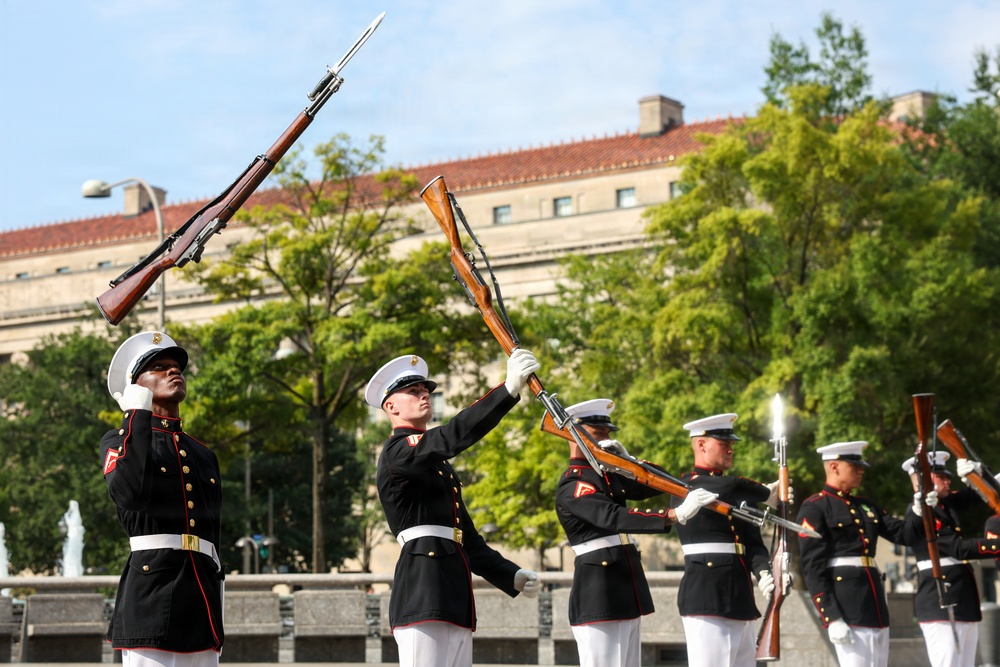 A group of U.S. Marine Corps ceremonial drill team members are performing a precision rifle drill in an outdoor urban setting. The Marines are dressed in formal dress blues uniforms with white covers, gloves, and belts, tossing and manipulating bayonet-tipped rifles in synchronized movements. The background features a large neoclassical government-style building and lush green trees, suggesting a Washington D.C. location.