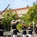A group of U.S. Marine Corps ceremonial drill team members are performing a precision rifle drill in an outdoor urban setting. The Marines are dressed in formal dress blues uniforms with white covers, gloves, and belts, tossing and manipulating bayonet-tipped rifles in synchronized movements. The background features a large neoclassical government-style building and lush green trees, suggesting a Washington D.C. location.
