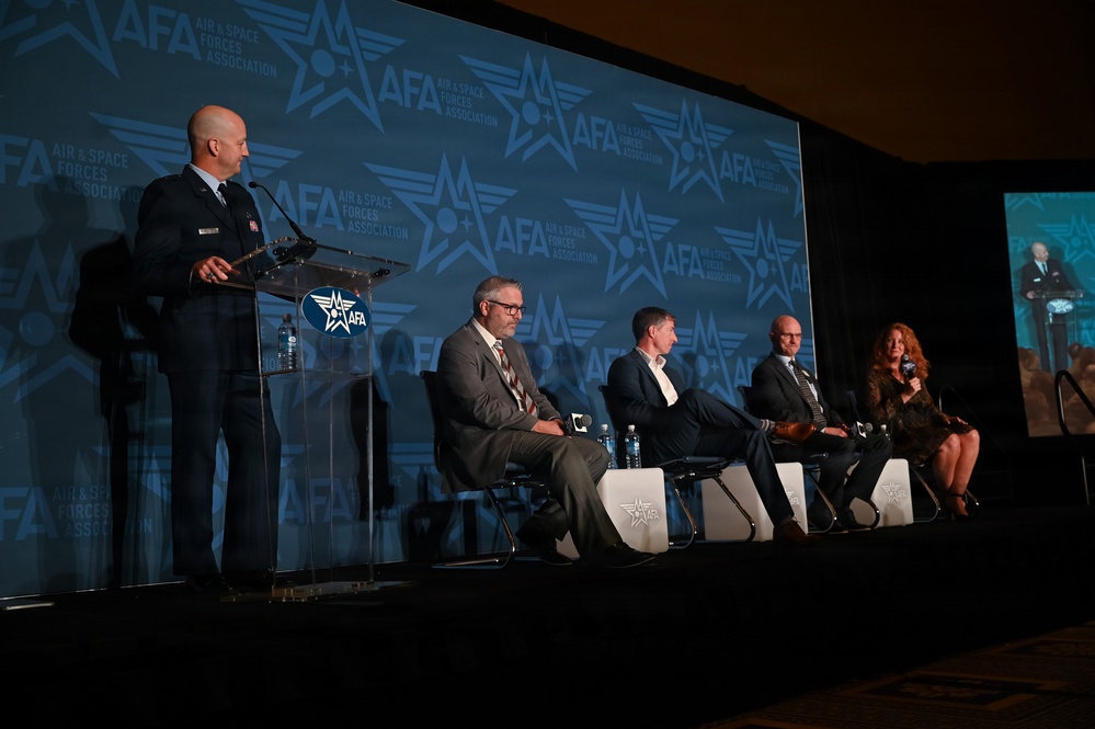 A panel discussion is taking place at an AFA (Air &amp; Space Forces Association) conference or symposium. A uniformed military officer stands at a podium on the left while four civilian panelists are seated in chairs on stage. The backdrop prominently displays the AFA logo and branding repeated across a large blue screen.