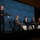 DAF Battle Network: Why is this Crucial? AFA PanelNext-Generation Battle Management - AFA Panel A panel discussion is taking place at an AFA (Air & Space Forces Association) conference or symposium. A uniformed military officer stands at a podium on the left while four civilian panelists are seated in chairs on stage. The backdrop prominently displays the AFA logo and branding repeated across a large blue screen.