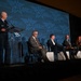 A panel discussion is taking place at an AFA (Air &amp; Space Forces Association) conference or symposium. A uniformed military officer stands at a podium on the left while four civilian panelists are seated in chairs on stage. The backdrop prominently displays the AFA logo and branding repeated across a large blue screen.