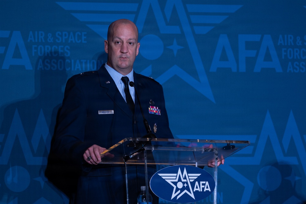 A uniformed U.S. Air Force or Space Force officer is speaking at a podium during an Air &amp; Space Forces Association (AFA) event. The officer is wearing a dark blue service dress uniform with multiple ribbons and decorations. The backdrop prominently displays the AFA logo repeated across a blue-toned background.