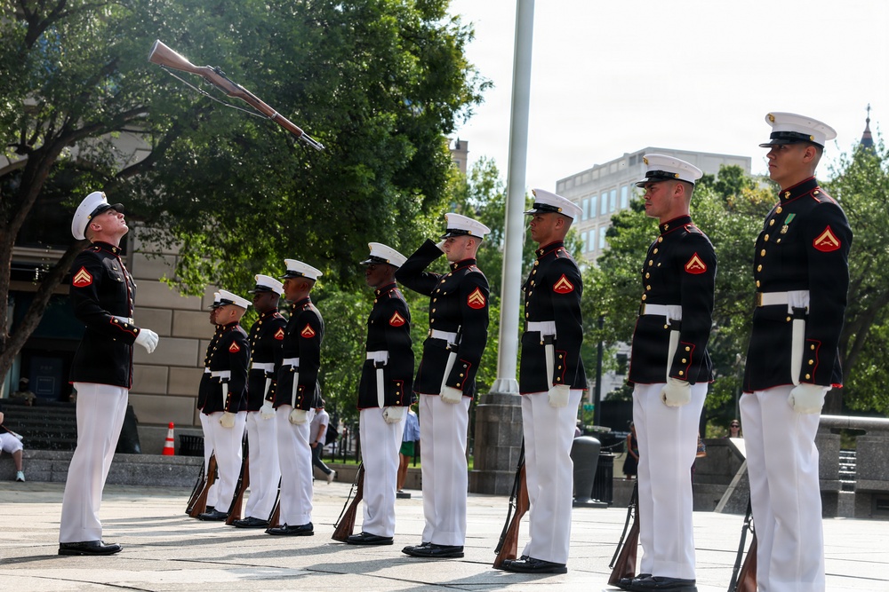 A U.S. Marine Corps Silent Drill Platoon performs a precision rifle exhibition in an outdoor urban plaza. Marines in full dress blues — black tunics with red trim, white trousers, and white covers — are lined up in formation while one Marine tosses a rifle into the air. The setting appears to be a public ceremonial event, likely in Washington, D.C., based on the surrounding architecture and trees.