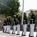 A U.S. Marine Corps Silent Drill Platoon performs a precision rifle exhibition in an outdoor urban plaza. Marines in full dress blues — black tunics with red trim, white trousers, and white covers — are lined up in formation while one Marine tosses a rifle into the air. The setting appears to be a public ceremonial event, likely in Washington, D.C., based on the surrounding architecture and trees.