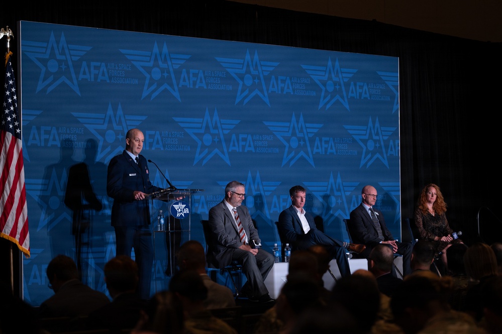 A panel discussion or presentation is taking place at an AFA (Air &amp; Space Forces Association) event. A uniformed military officer stands at a podium on the left, while four civilian panelists — three men and one woman — are seated in chairs on the stage. An American flag is visible on the far left, and the AFA branded backdrop dominates the background.