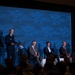 A panel discussion or presentation is taking place at an AFA (Air &amp; Space Forces Association) event. A uniformed military officer stands at a podium on the left, while four civilian panelists — three men and one woman — are seated in chairs on the stage. An American flag is visible on the far left, and the AFA branded backdrop dominates the background.
