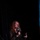 Next-Generation Battle Management - AFA Panel A woman with curly red hair is seated on a panel stage speaking into a microphone branded with the AFA (Air Force Association) logo. She is wearing a dark floral-patterned dress and gesturing with her free hand, suggesting active engagement in a discussion. The event appears to be an AFA conference or symposium, indicated by the AFA branding visible on both the microphone and a white podium/stand in the foreground.