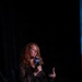 A woman with curly red hair is seated on a panel stage speaking into a microphone branded with the AFA (Air Force Association) logo. She is wearing a dark floral-patterned dress and gesturing with her free hand, suggesting active engagement in a discussion. The event appears to be an AFA conference or symposium, indicated by the AFA branding visible on both the microphone and a white podium/stand in the foreground.