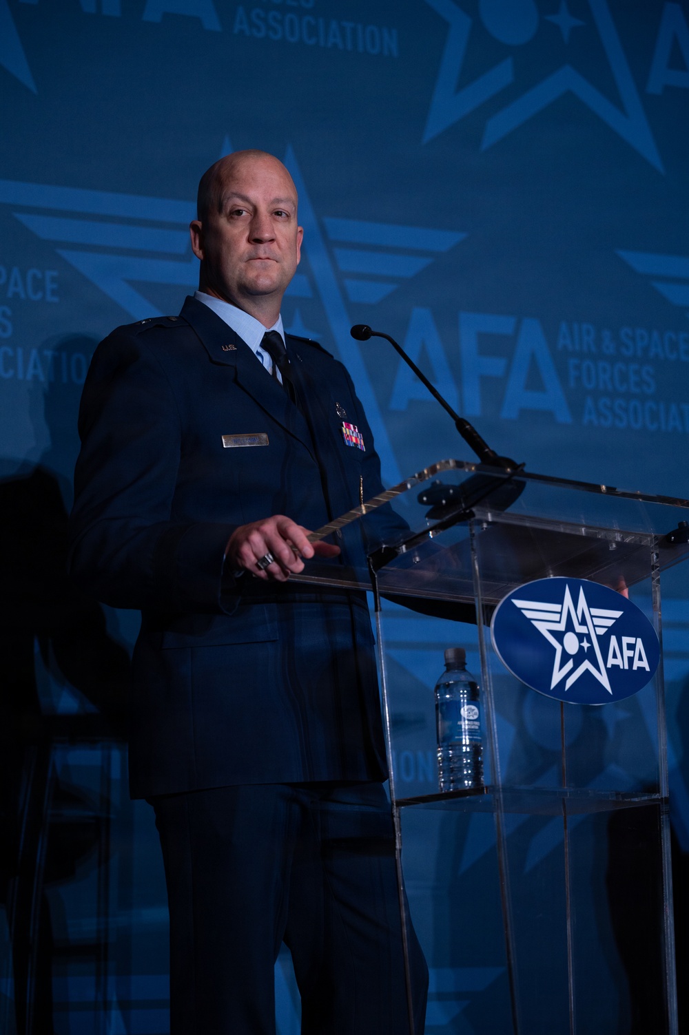 A bald male military officer in a dark blue dress uniform stands at a clear acrylic podium speaking at an Air &amp; Space Forces Association (AFA) event. He is wearing multiple service ribbons and medals on his chest, with a name tag visible on his left breast. The background features a projected AFA logo and branding in blue tones.