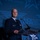 Next-Generation Battle Management - AFA Panel A bald male military officer in a dark blue dress uniform stands at a clear acrylic podium speaking at an Air & Space Forces Association (AFA) event. He is wearing multiple service ribbons and medals on his chest, with a name tag visible on his left breast. The background features a projected AFA logo and branding in blue tones.