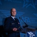 A bald male military officer in a dark blue dress uniform stands at a clear acrylic podium speaking at an Air &amp; Space Forces Association (AFA) event. He is wearing multiple service ribbons and medals on his chest, with a name tag visible on his left breast. The background features a projected AFA logo and branding in blue tones.