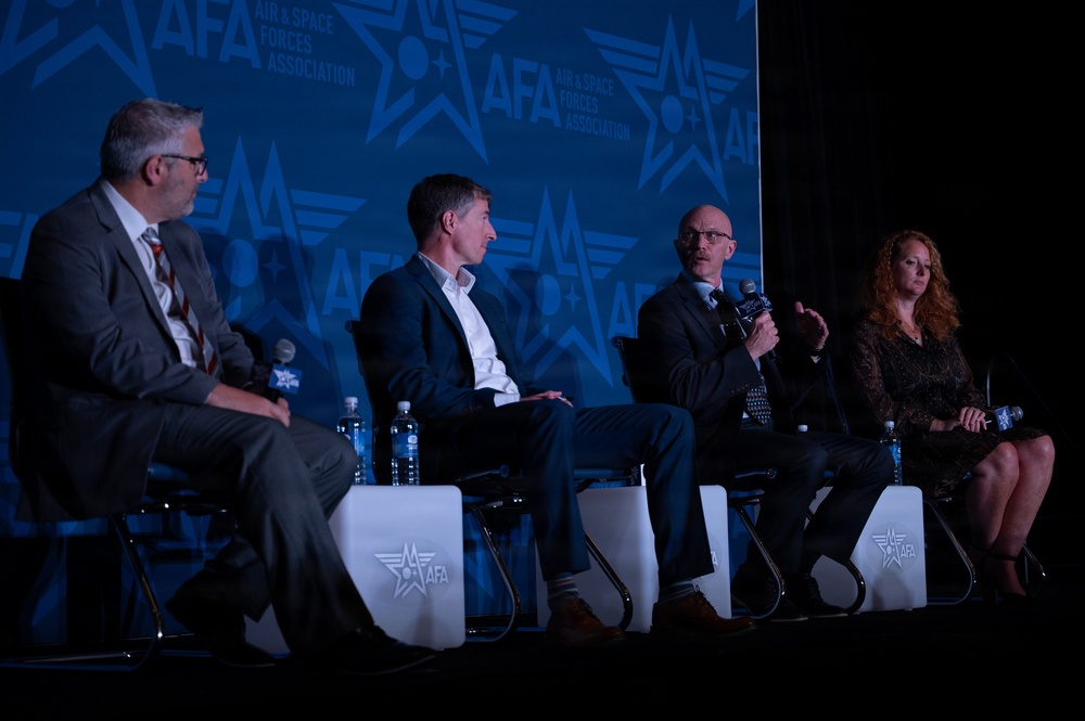 Four panelists are seated on a stage at an AFA (Air &amp; Space Forces Association) event, engaged in a panel discussion. One panelist is actively speaking into a microphone while the others listen. The backdrop prominently displays the AFA logo and branding in blue tones.