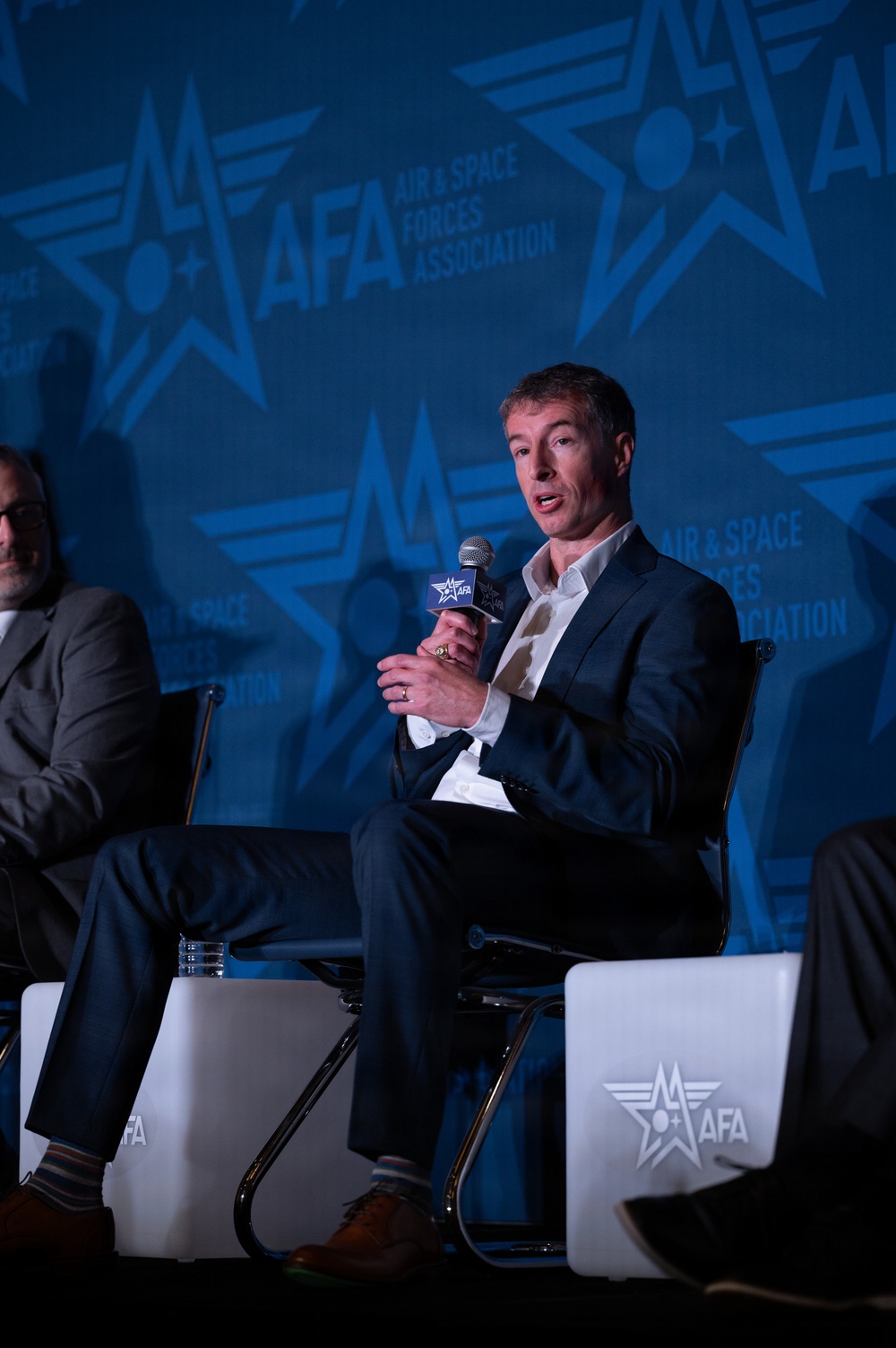 A man in a navy blue suit with an open-collar white shirt is seated on a panel stage, speaking into an AFA-branded microphone. The event is hosted by the Air &amp; Space Forces Association (AFA), as indicated by the prominent branded backdrop and podium signage. At least one other panelist is partially visible to his left.