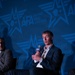 A man in a navy blue suit with an open-collar white shirt is seated on a panel stage, speaking into an AFA-branded microphone. The event is hosted by the Air &amp; Space Forces Association (AFA), as indicated by the prominent branded backdrop and podium signage. At least one other panelist is partially visible to his left.