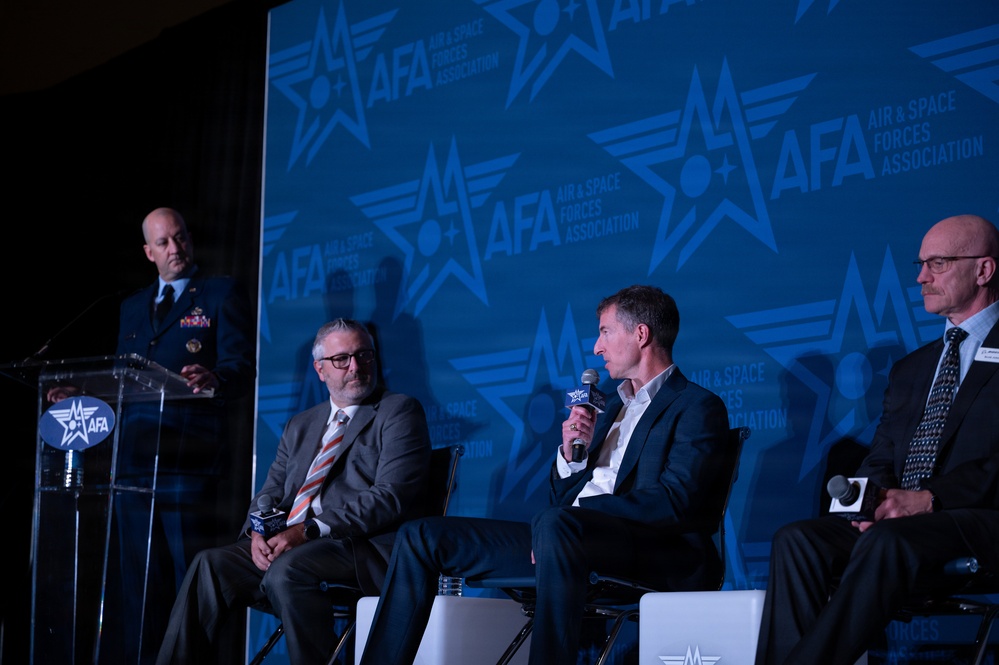 A panel discussion is taking place at an Air &amp; Space Forces Association (AFA) event. Four individuals are on stage: a uniformed military officer standing at a podium on the left, and three civilians seated in white chairs. One of the seated panelists is speaking into a microphone against a large blue AFA-branded backdrop.