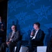 A panel discussion is taking place at an Air &amp; Space Forces Association (AFA) event. Four individuals are on stage: a uniformed military officer standing at a podium on the left, and three civilians seated in white chairs. One of the seated panelists is speaking into a microphone against a large blue AFA-branded backdrop.