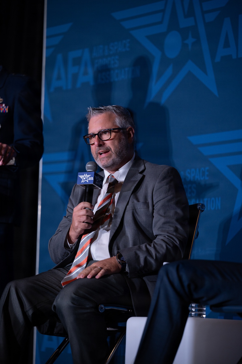 A middle-aged man with gray hair and a beard is seated in a chair, speaking into a microphone branded with the AFA (Air Force Association) logo. He is dressed in a gray suit with a white shirt and an orange-and-white striped tie. The event backdrop displays the AFA 'Air &amp; Space Forces Association' branding prominently.