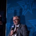 A middle-aged man with gray hair and a beard is seated in a chair, speaking into a microphone branded with the AFA (Air Force Association) logo. He is dressed in a gray suit with a white shirt and an orange-and-white striped tie. The event backdrop displays the AFA 'Air &amp; Space Forces Association' branding prominently.