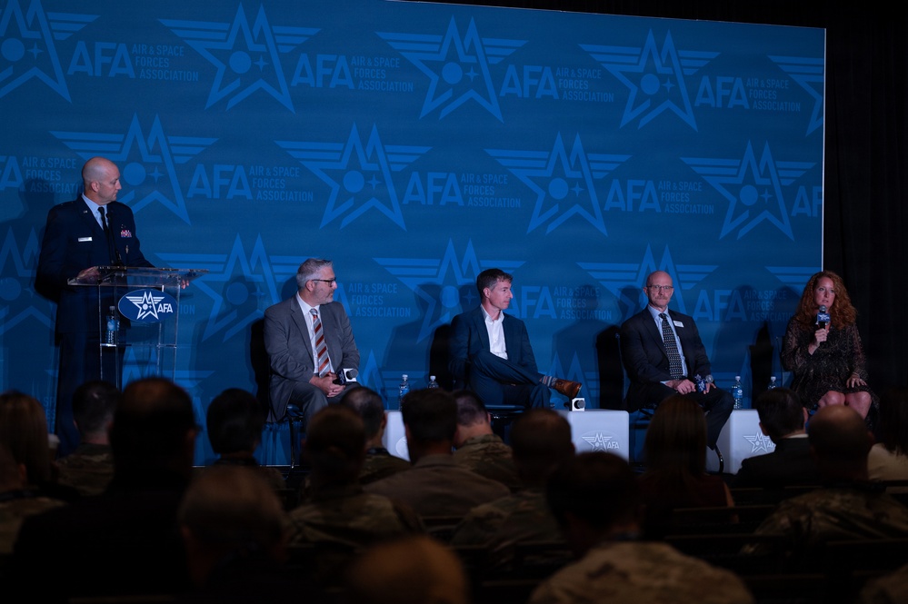 A panel discussion is taking place at an AFA (Air &amp; Space Forces Association) event or conference. A uniformed military officer stands at a podium on the left while four civilian panelists — three men and one woman — are seated on stage. A large audience, including military personnel in uniform, is visible in the foreground.
