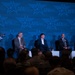 A panel discussion is taking place at an AFA (Air &amp; Space Forces Association) event or conference. A uniformed military officer stands at a podium on the left while four civilian panelists — three men and one woman — are seated on stage. A large audience, including military personnel in uniform, is visible in the foreground.