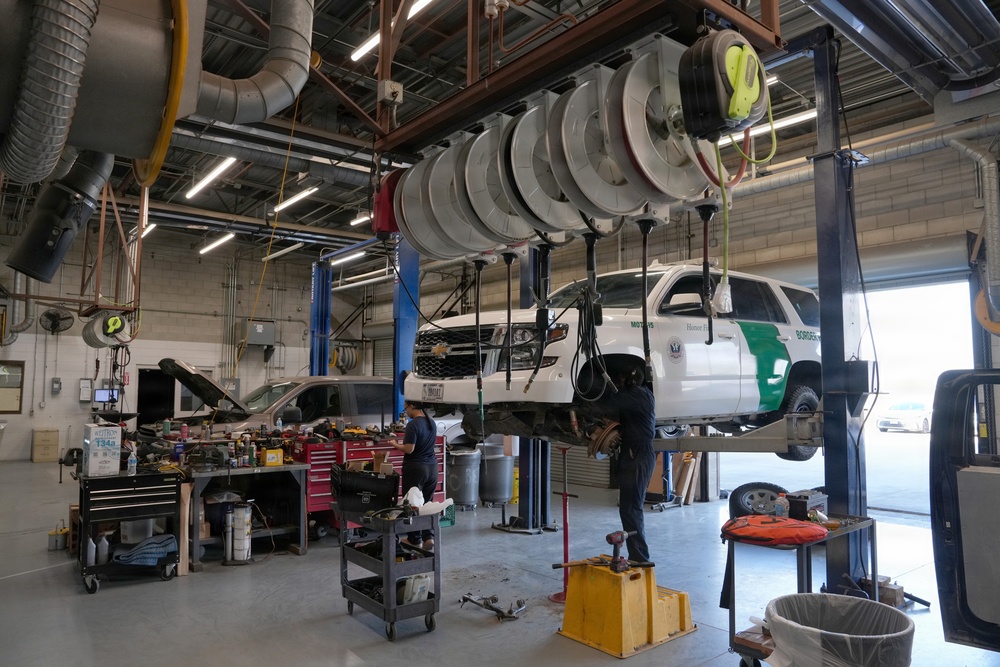 A professional automotive repair shop with two mechanics working on vehicles. A white U.S. Border Patrol SUV is elevated on a hydraulic lift while a second vehicle with its hood open is being serviced nearby. The facility appears to be a government fleet maintenance garage equipped with industrial overhead hose reels and professional tooling.