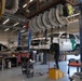 A professional automotive repair shop with two mechanics working on vehicles. A white U.S. Border Patrol SUV is elevated on a hydraulic lift while a second vehicle with its hood open is being serviced nearby. The facility appears to be a government fleet maintenance garage equipped with industrial overhead hose reels and professional tooling.