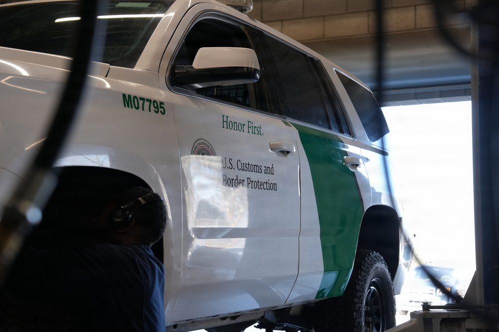 A U.S. Customs and Border Protection (CBP) vehicle, unit number M07795, is elevated on a lift inside what appears to be a maintenance or repair facility. The white SUV bears the agency's official markings including the motto 'Honor First' and the CBP seal. A person, likely a mechanic, is visible beneath or beside the vehicle performing service work.