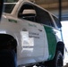 A U.S. Customs and Border Protection (CBP) vehicle, unit number M07795, is elevated on a lift inside what appears to be a maintenance or repair facility. The white SUV bears the agency's official markings including the motto 'Honor First' and the CBP seal. A person, likely a mechanic, is visible beneath or beside the vehicle performing service work.