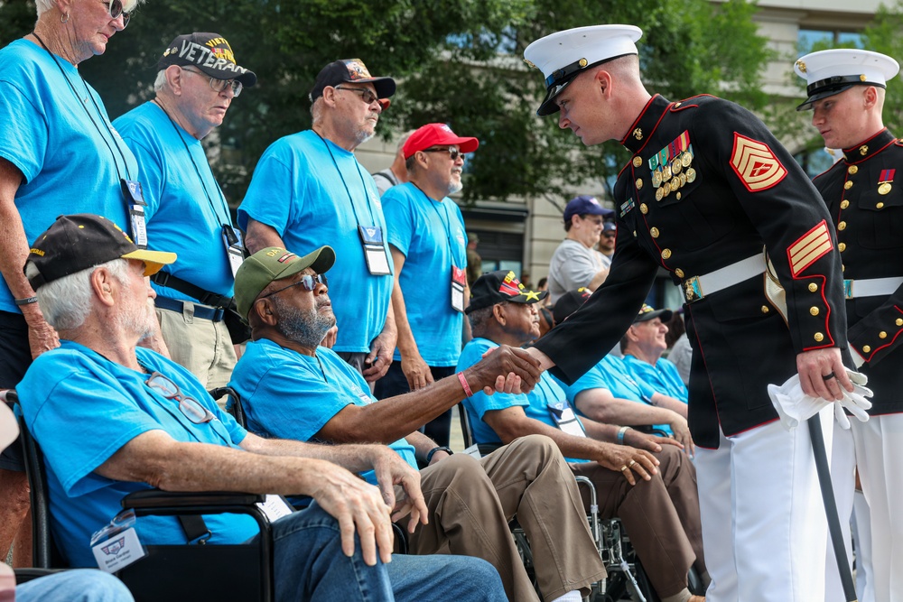 A U.S. Marine in dress blues uniform is shaking hands with a veteran seated in a wheelchair, who is wearing a green cap and blue t-shirt. The group of veterans, all wearing matching light blue t-shirts and lanyards with badges, appear to be participating in an organized honor event, likely a veterans recognition ceremony. Two Marines in formal dress uniforms are greeting the assembled veterans outdoors.