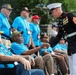 A U.S. Marine in dress blues uniform is shaking hands with a veteran seated in a wheelchair, who is wearing a green cap and blue t-shirt. The group of veterans, all wearing matching light blue t-shirts and lanyards with badges, appear to be participating in an organized honor event, likely a veterans recognition ceremony. Two Marines in formal dress uniforms are greeting the assembled veterans outdoors.