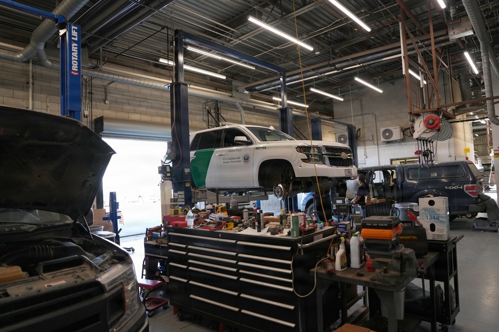The image shows a government vehicle maintenance facility with a U.S. Customs and Border Protection (CBP) SUV elevated on a Rotary brand two-post lift. The shop is well-equipped with tool chests, various automotive tools, and multiple vehicles being serviced simultaneously. A mechanic is visible working in the background near the lifted CBP vehicle.