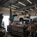 The image shows a government vehicle maintenance facility with a U.S. Customs and Border Protection (CBP) SUV elevated on a Rotary brand two-post lift. The shop is well-equipped with tool chests, various automotive tools, and multiple vehicles being serviced simultaneously. A mechanic is visible working in the background near the lifted CBP vehicle.