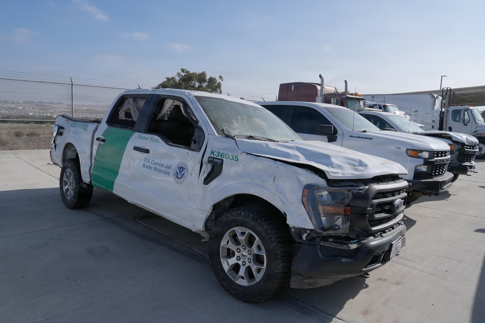 A heavily damaged U.S. Customs and Border Protection (CBP) Ford F-150 pickup truck, unit number K36035, is parked in what appears to be an impound or vehicle storage lot. The vehicle shows significant collision damage including a crushed roof, damaged hood, missing or broken windows, and body panel deformation. Several other vehicles are visible in the background, including a Chevrolet Silverado and a semi-truck.