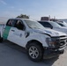 A heavily damaged U.S. Customs and Border Protection (CBP) Ford F-150 pickup truck, unit number K36035, is parked in what appears to be an impound or vehicle storage lot. The vehicle shows significant collision damage including a crushed roof, damaged hood, missing or broken windows, and body panel deformation. Several other vehicles are visible in the background, including a Chevrolet Silverado and a semi-truck.