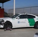 A damaged U.S. Customs and Border Protection (Border Patrol) vehicle, unit number E94096, is shown parked on a concrete surface. The white Dodge Charger patrol car has sustained significant front-end collision damage, including a destroyed front bumper, damaged hood, and a missing or flat front tire supported by a rolling dolly. The vehicle appears to be in a secured facility surrounded by chain-link fencing topped with razor wire.