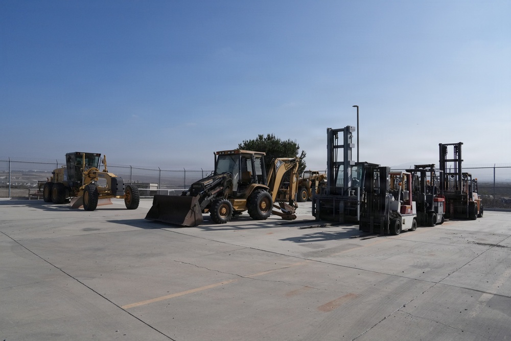 A collection of heavy construction and material handling equipment is parked on a concrete surface, likely a staging or storage yard. The equipment includes motor graders, a backhoe loader, and multiple forklifts of varying sizes. The setting appears to be an industrial or military facility enclosed by a chain-link fence.