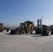 A collection of heavy construction and material handling equipment is parked on a concrete surface, likely a staging or storage yard. The equipment includes motor graders, a backhoe loader, and multiple forklifts of varying sizes. The setting appears to be an industrial or military facility enclosed by a chain-link fence.