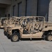 Multiple military utility terrain vehicles (UTVs) are parked in a row alongside a concrete block building with roll-up garage doors. The vehicles are painted in a uniform tan/desert sand color consistent with military specifications. They appear to be open-frame, multi-seat tactical vehicles with roll cages, likely used for special operations or light reconnaissance missions.
