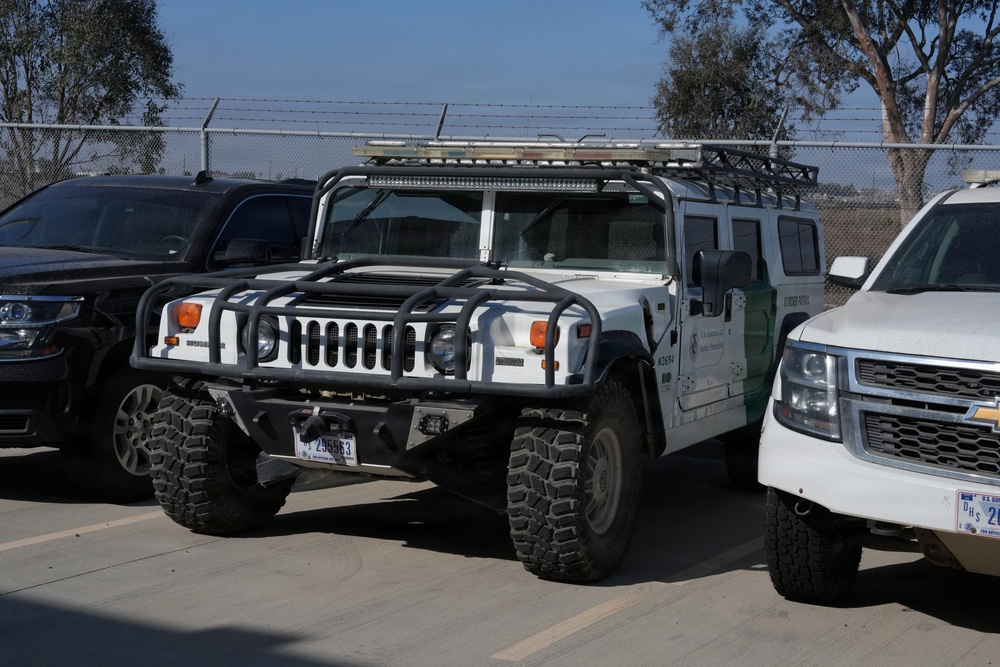 The image shows a white Hummer H1 vehicle equipped with off-road modifications, parked in a secured lot alongside other vehicles. The Hummer bears markings consistent with U.S. Customs and Border Protection (CBP), including a green stripe and agency insignia on the door. The setting appears to be a government or law enforcement facility, indicated by the chain-link fence with barbed wire in the background.