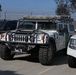 The image shows a white Hummer H1 vehicle equipped with off-road modifications, parked in a secured lot alongside other vehicles. The Hummer bears markings consistent with U.S. Customs and Border Protection (CBP), including a green stripe and agency insignia on the door. The setting appears to be a government or law enforcement facility, indicated by the chain-link fence with barbed wire in the background.